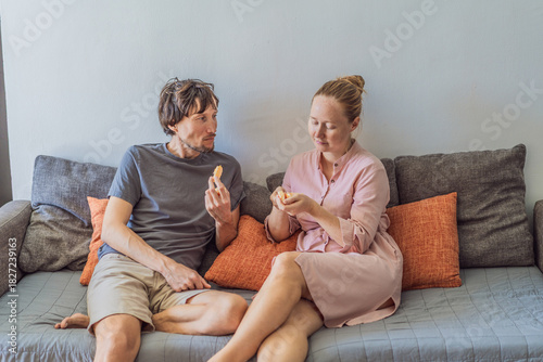 Man and woman sitting together on a sofa eating an orange, sharing a relaxed moment at home in a cozy living room. Concept of family life, togetherness, healthy snack, everyday lifestyle and real
