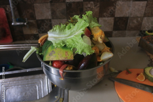 A close-up shot of a stainless steel bowl filled with various organic food scraps and kitchen waste, including lettuce leaves, corn cobs, tomato ends, watermelon rinds, and other vegetable and fruit p