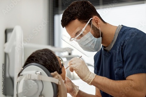 Photography Young adult male dentist wearing protective mask and face shield treating patien