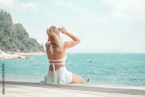 A young woman in a white bikini sits on the sea wall, hand in hair, gazing at the turquoise ocean with lush cliffs behind, capturing pure summer beach bliss. Tropical paradise, vacation vibe.