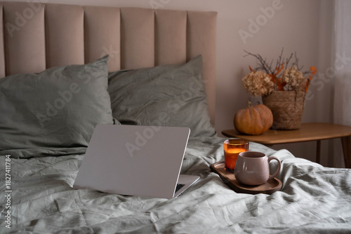 Cozy bedroom scene with a laptop on a bed, warm autumn decor, coffee and candle on a wooden tray, creating a calm and comfortable home atmosphere.