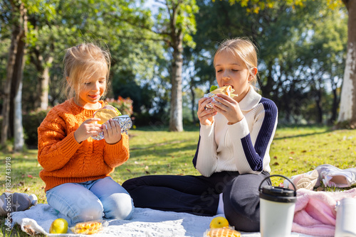 Two young girls enjoying sandwiches during a picnic in a sunny park