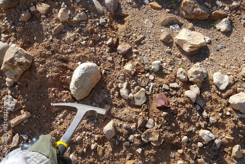 Geologist's gloved hand holds rock hammer above scattered stones and sandy soil at a sunny quarry site