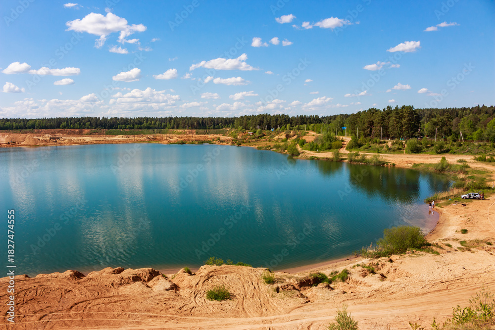 Obraz premium Stunning turquoise water in a sandy quarry lake surrounded by lush green forest under a bright blue sky with fluffy clouds