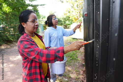 Two women are helping each other paint a black metal fence. The woman in the foreground is using a paintbrush, while the one behind her is using a paint roller.