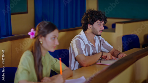 Indian young adult gen z busy pupil boy girl guys sitting on desk table hold pencil do study work exam notes class room. Smart male man female learn new task project day time inside college place