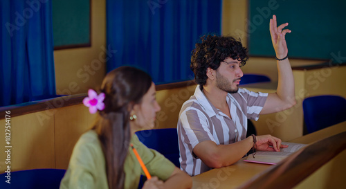 Indian young adult gen z busy boy man guy pair sitting desk table hold pencil paper do study work writing notes pupil female girl male raise hand talk share new idea day inside class room place