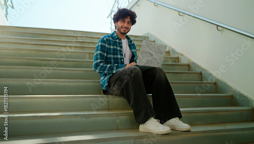 Indian young adult gen z male man use laptop look screen do study read email work check online e learning course break time. Happy pupil boy staring cam do casual pose enjoy new day outdoor place	