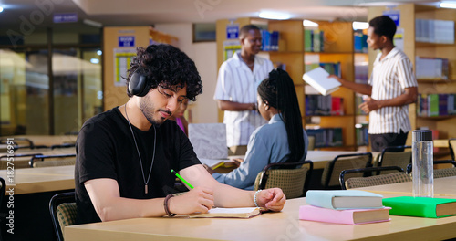 Indian Asian young adult gen z male man calm peace mind student sitting desk table listening music hold pencil write exam notes smart boy do self study read book prepare topics indoor library busy day