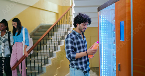 Indian happy young adult gen z smart male man use easy fast self service choose juice drink campus area. Lady female walk stairway thirsty boy take bottle enjoy break day time hallway