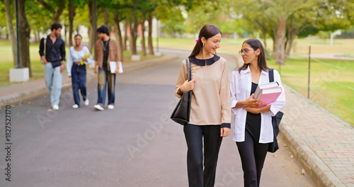 Indian happy young adult gen z lady female walk wear glass talk gossip share new day plan enjoy break time outdoor place. Teen pupil girl guys hold book speak funny joke have fun joy park