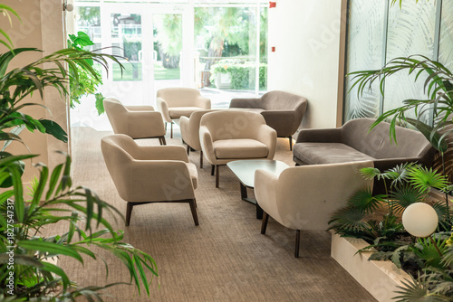 A modern lounge area with several light-colored armchairs arranged around a glass table. Green plants add a touch of nature to the space.