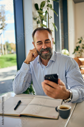 Confident Man At Coffee Shop Checks Smartphone While Reviewing Planner