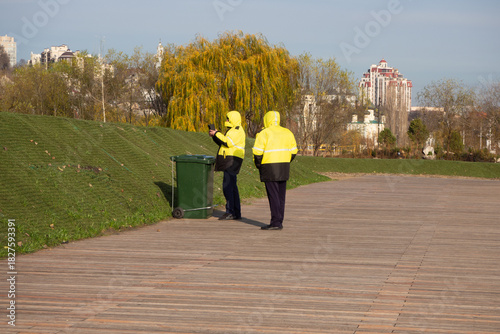 Two workers in yellow uniforms near green trash bin on wooden boardwalk. Public service, urban maintenance, waste management. Real photo