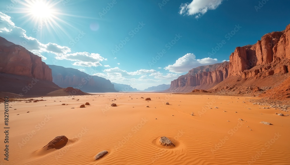Naklejka premium Expansive desert landscape with towering red rock cliffs on both sides of wide valley. Bright sun illuminates golden sandy terrain with ripples, scattered rocks. Clear blue sky, white clouds over