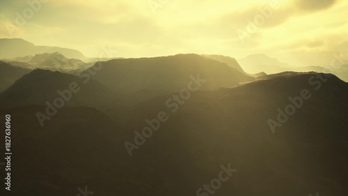 Silhouetted mountain ranges rise dramatically against a backdrop of warm golden light as dusk approaches. Soft shadows blend with vibrant hues over the serene landscape, evoking tranquility.