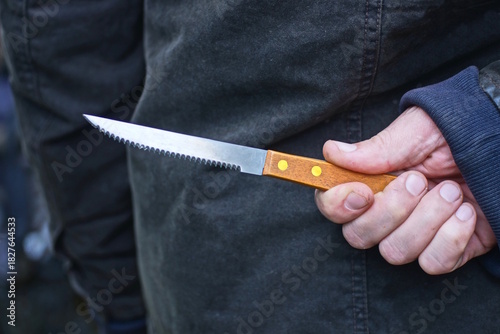 hand of a male criminal in black clothes and a gray brown knife behind his back on the street
