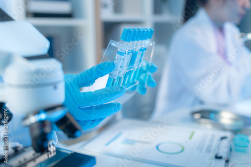 Two female scientists in white lab coats working together in a bright laboratory with microscopes, glassware, and blue test tubes, representing teamwork and modern scientific research environment.