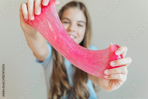 Close-up of hands stretching bright pink slime towards the camera, with a smiling girl in the background. Sensory toy