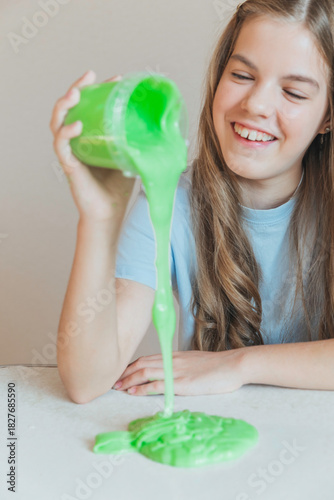 Smiling girl pouring vibrant green slime from a container onto the table. Trendy DIY and fun sensory play activity
