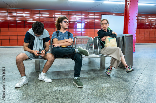Berlin, Germany, August 10, 2023. A subway station in Frankfurt  Allee, characterized by its red and orange tiled walls. On a bench, two young men and a middle-aged woman wait for the train to pass.