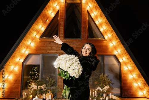 Woman holding white roses waving in front of a wooden house with lights