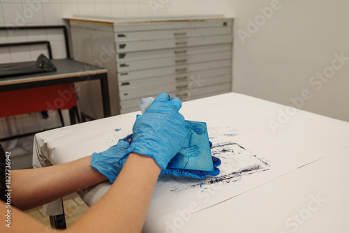 Close up of hands wearing blue protective gloves wiping a blue printing plate on a white table in an art studio with a flat file cabinet in the background.