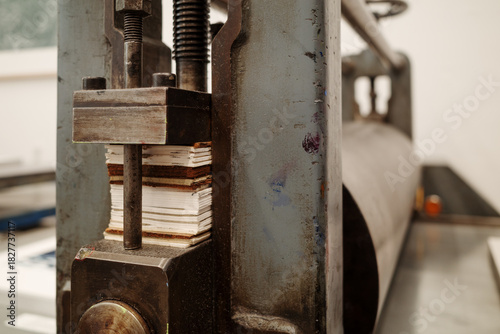 A detailed view of a heavy metal printing press in a workshop setting. The image focuses on the threaded screw and a stack of shims used to adjust the pressure of the roller.
