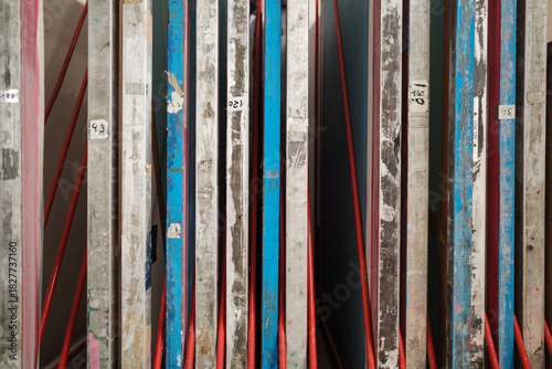 A close-up side view of used screen printing frames stored vertically in a red metal rack within an art studio. The aluminum and wooden frames are stained with dried blue and pink ink. 