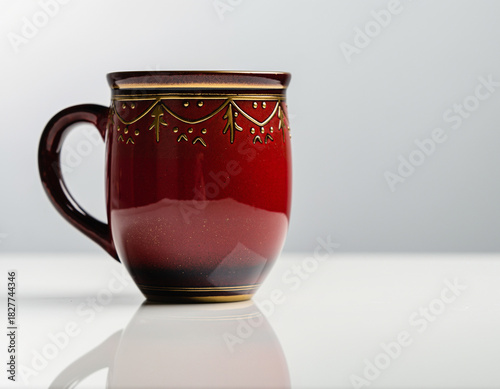 Red ceramic mug sits on a white table against a light grey backdrop, showcasing its intricate gold trim and glossy surface in a minimalist setting.