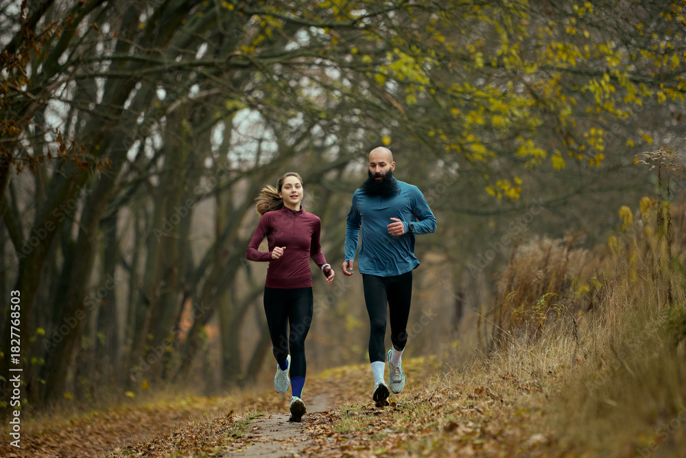 Naklejka premium Active runners moving forward on forest trail in crisp autumn season. Concept of outdoor vitality, functional fitness, lifestyle motivation, healthy habits and seasonal wellness engagement.
