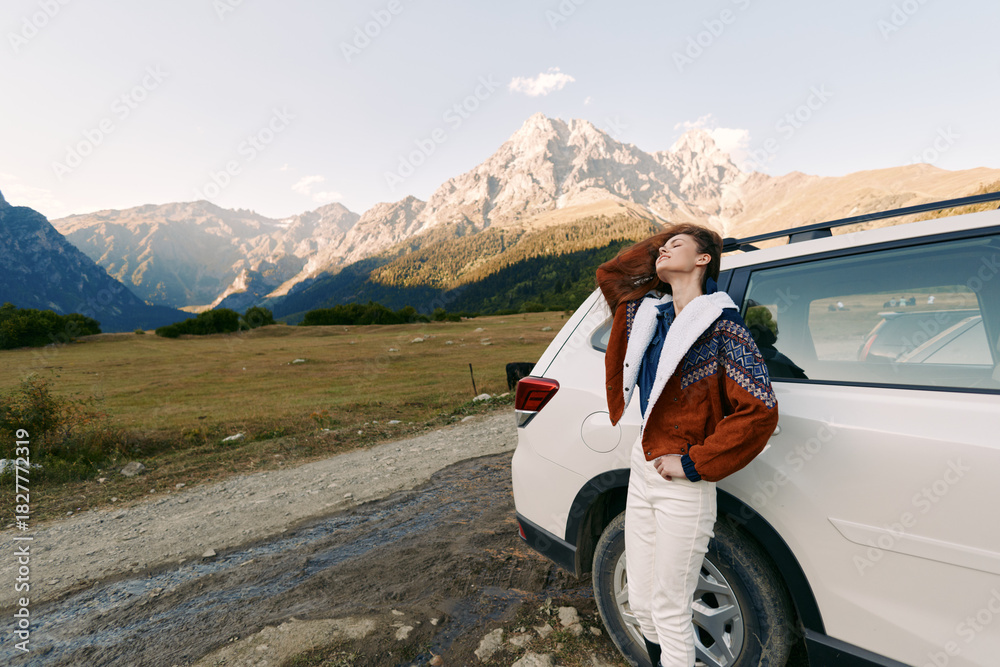 Obraz premium Woman by car at roadside with mountains in background, enjoying travel and nature during a scenic road trip, relaxed pose leaning on white SUV at sunset landscape.