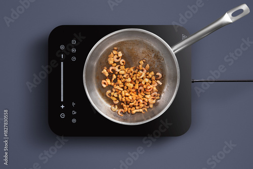 Shrimp are sizzling in a pan on an induction stove. A modern kitchen setting provides a sleek backdrop for the cooking activity. The focus is on the shrimp being cooked.