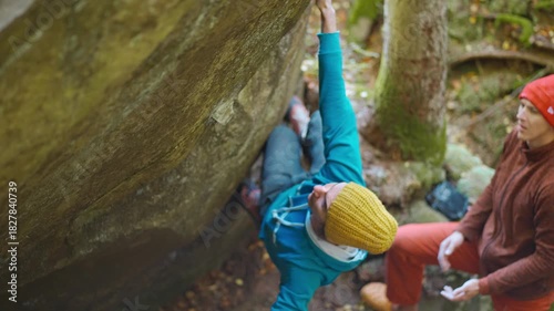 slow motion rock climbing outdoors in autumn forest. friends rock climbers climbs bouldering belaying each other. man in bright blue hoodie and yellow knitted cap hanging on edge of overhanging cliff