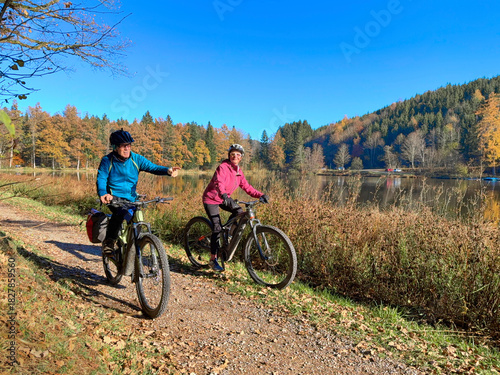 nice active senior couple riding their electric mountain bike at the pond of Hengelesweiher near city of Isny in the Swabian  Allgaeu on warm autumn day, Baden-Wuerttemberg, Germany