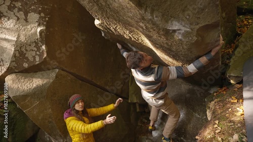 man rock climber climbs on overhanging boulder by it edges using wide hands grips, friends belay him. climbing outdoor in autumn forest on natural rocks. healthy active lifestyle, training outdoors.
