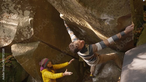 strong adult male climber climbs on overhanging boulder by it edges using wide hands grips, woman belay him. climbing outdoor in autumn forest. healthy active lifestyle, training outdoors.