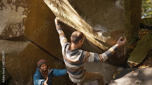 strong adult male climber climbs on overhanging boulder by it edges using wide hands grips, woman belay him. climbing outdoor in autumn forest. healthy active lifestyle, training outdoors.