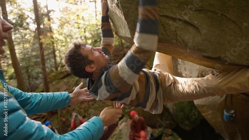 man rock climber climbs on overhanging boulder by it edges using wide hands grips, friends belay him. climbing outdoor in autumn forest on natural rocks. healthy active lifestyle, training outdoors.