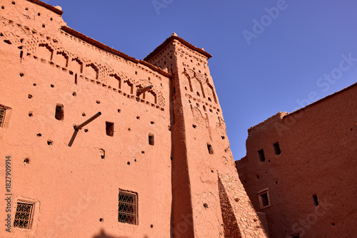 The earthen buildings of the historic Ksar of Ait ben Haddou rise against a clear blue sky. The traditional architecture shows detail and texture.