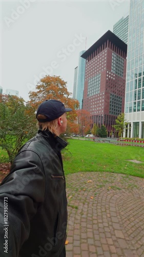 A man walks through a park in a city, surrounded by modern office buildings and autumn foliage