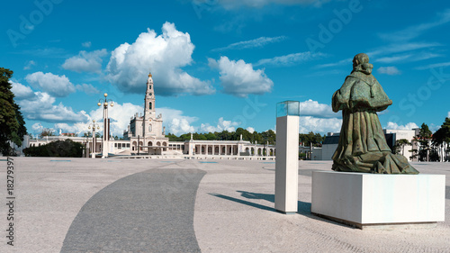 Bronze statue of Pope Paul VI, praying, kneeling with weathered green patina stands before Basilica of Our Lady of the Rosary at Sanctuary of Fátima, Portugal, iconic religious monument under blue sky