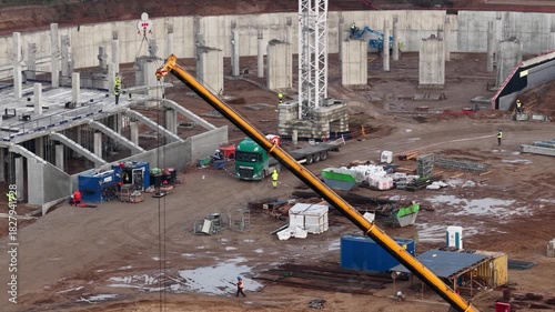 Comprehensive construction scene showing a partially built stadium structure, foundation grid, heavy machinery, materials, and active workforce on muddy terrain.