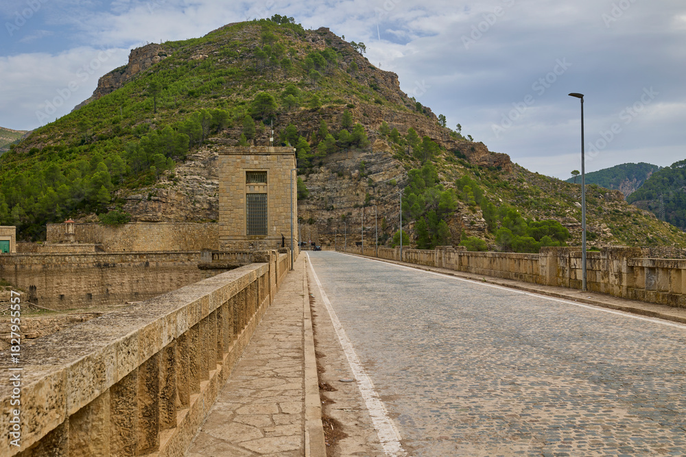 Fototapeta premium Scenic road built along the top of a massive stone dam, surrounded by mountain hills and greenery in Spain.