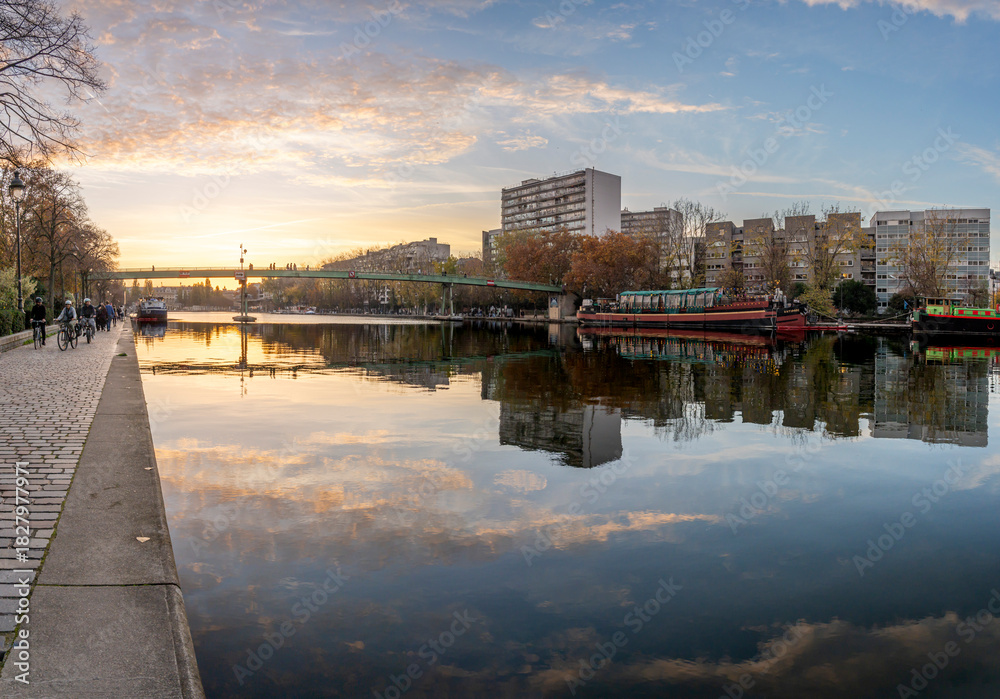 Naklejka premium Paris, France - 11 08 2025: Ourcq Canal. Panoramic View of the Villette Basin, Moselle Footbridge and buildings behind reflecting on the water at sunset and golden hour
