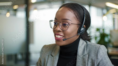 A confident young african american woman in a grey blazer and headset smiles and speaks articulately, focused left in a modern corporate office, blurred bokeh background. Professional virtual