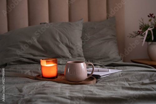 Cozy bedroom scene with a lit candle, warm mug, and notebook on a wooden tray placed on a bed with soft green bedding.