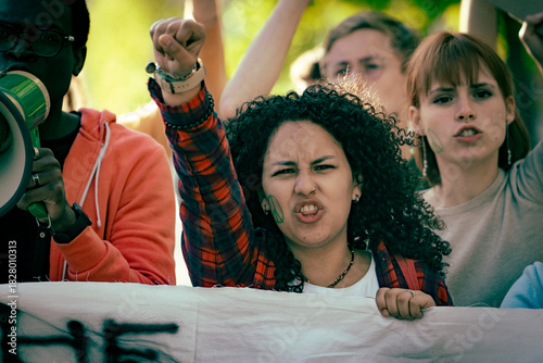 Portrait of a young female activist shouting and raising her fist during a protest outdoors, surrounded by a diverse group of demonstrators taking part in a social movement.