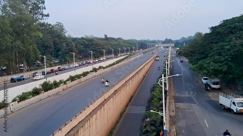 A wide view of a clean urban road with parallel lanes, light traffic, and greenery on both sides. The scene captures early morning or evening city movement with vehicles, trees, and well-maintained