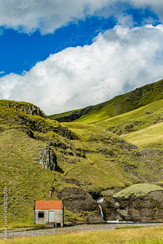 A rust-roofed refuge rests beside a cascading stream in Southern Iceland’s mossy hills under a patchwork sky
